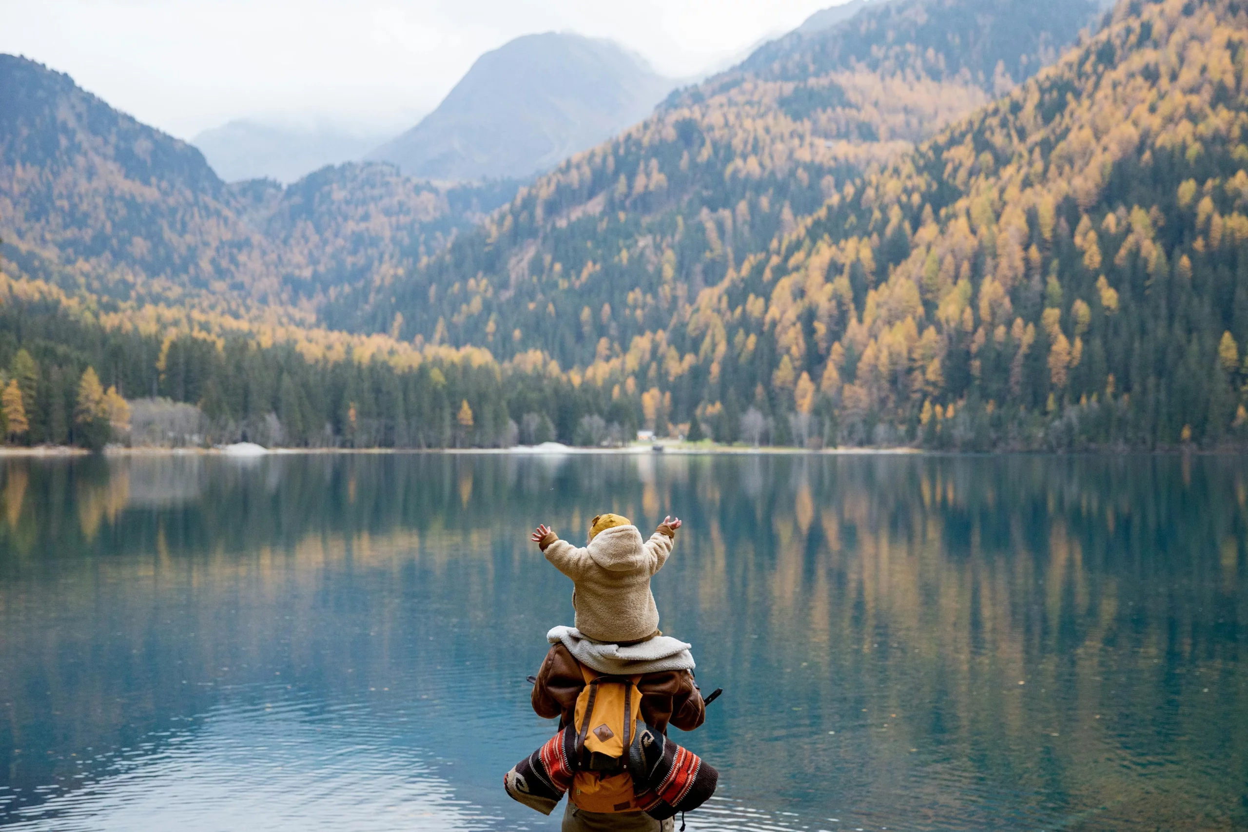 Voyageur assis au bord d’un lac de montagne avec une valise, symbole d’un voyage sur mesure haut de gamme en famille sous le soleil.